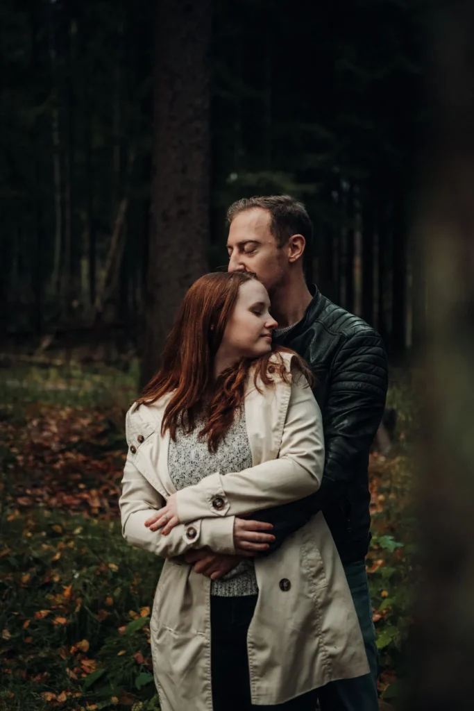 Angehendes Brautpaar steht romantisch im Wald, der Bräutigam umschließt die Braut von hinten mit seinen Armen, beide genießen den Moment voller Liebe.